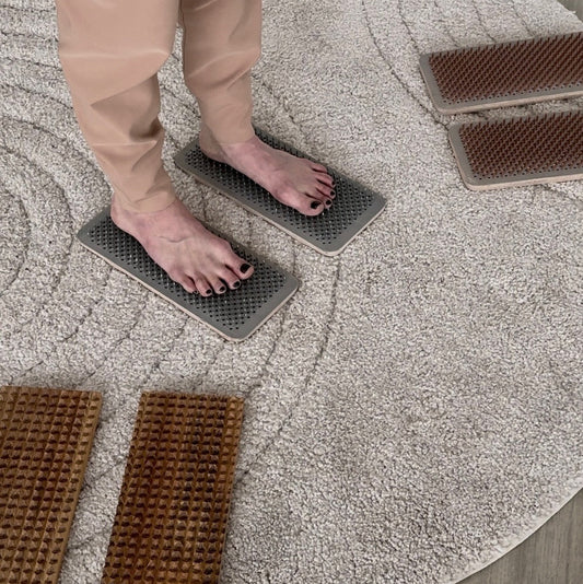 A person wearing beige pants stands barefoot on two gray textured foot mats placed on a round light rug over a wood floor. Two brown massage-like mats are also visible nearby.
