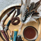 A styled flat-lay of a bohemian self-care and spiritual tray containing a feather wand, wooden beads, a bowl of brown liquid, crystals, and other handmade accessories on a round gold tray.