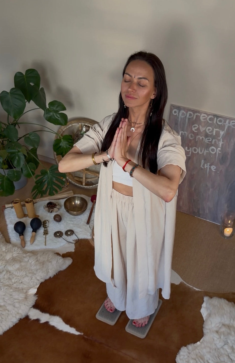 A woman in a light beige two-piece outfit stands on the nails in a room with her hands together in a prayer pose, surrounded by boho decor including a plant, candles, crystals, and a cowhide rug.