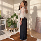 A woman in a bohemian-style outfit consisting of a white crochet shrug and a long black pleated maxi skirt stands on a scale in a sunlit room with plants and candles. Another woman is partially visible behind her.