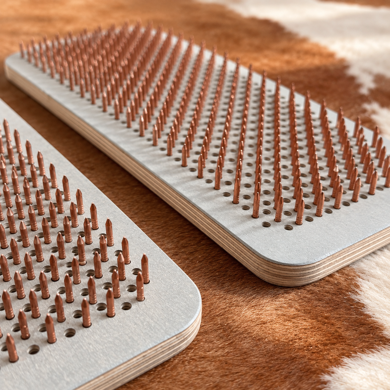 Two rectangular metal nail guns with rows of pointed metal nails, shown resting on a brown and white cowhide rug.